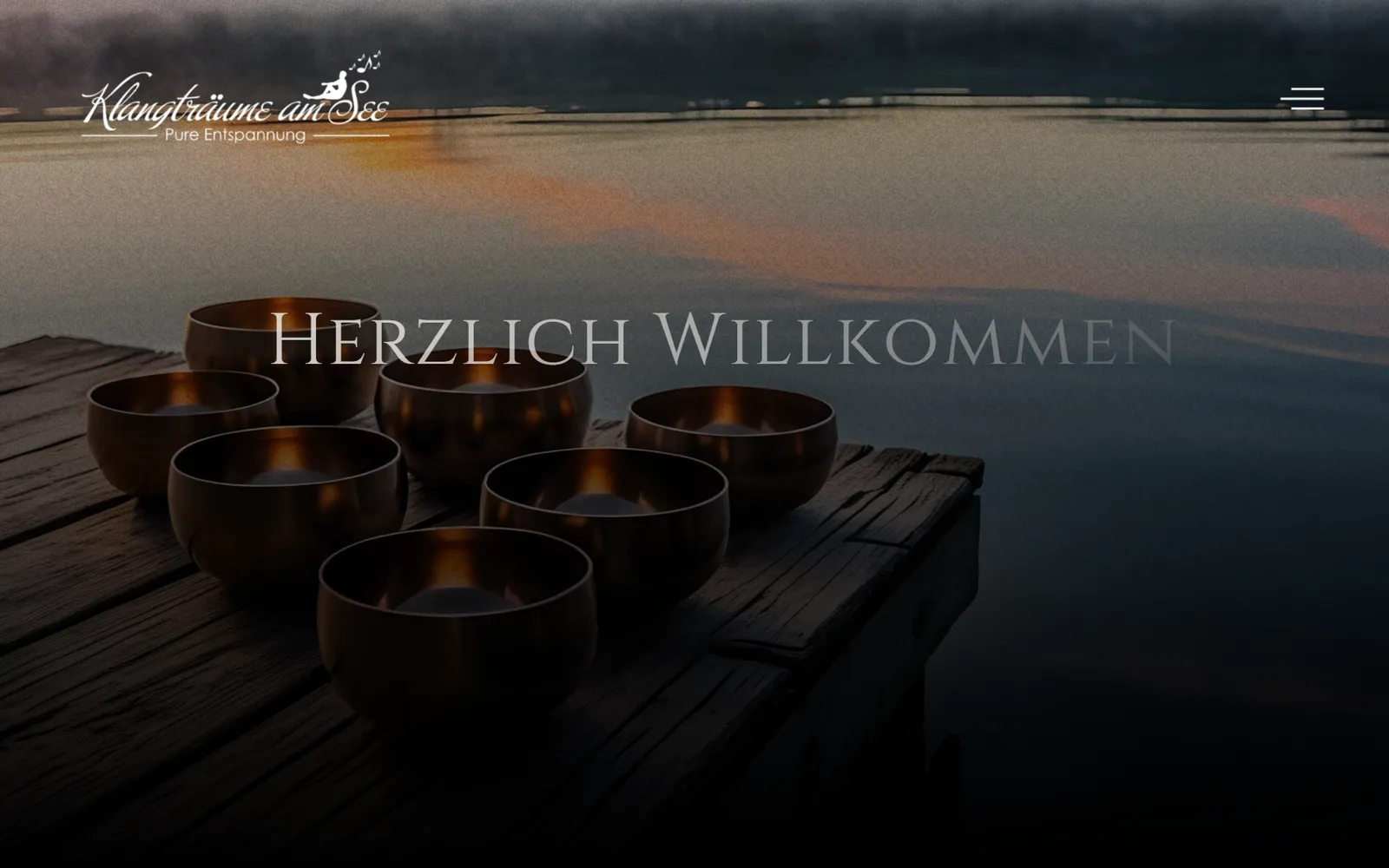 Startseite der Website von Klangträume am See: Klangschalen mit Kerzenlicht auf einem Steg am See in der Dämmerung, Überschrift Herzlich Willkommen